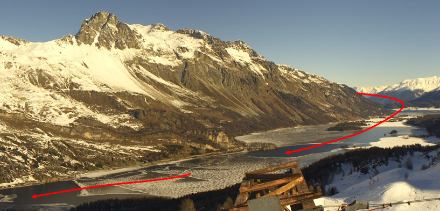 Image actuelle du lac de Sils avec vue vers le nord. En bas à gauche ainsi qu'au milieu du lac, on peut encore reconnaître les dernières surfaces ouvertes du lac. La partie nord, légèrement protégée du vent, entre Plaun da Lej et Sils-Baselagia, est déjà gelée depuis longtemps. Les flèches rouges indiquent la direction du courant du vent du nord.