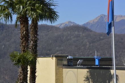 La couleur du détecteur de grêle bleu est en harmonie avec le drapeau et le ciel bleu du canton méridional suisse. Mais il peut aussi grêler au Tessin. Le détecteur de grêle sur le toit de la maison et le radar météorologique sur le Monte Lema en arrière-plan sont prêts à détecter la grêle 24 heures sur 24. 
