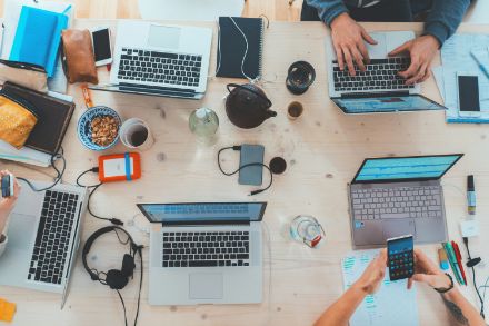 La photo montre une grande table en bois avec plusieurs ordinateurs portables, smartphones et documents de travail, tandis que des personnes travaillent et interagissent.
