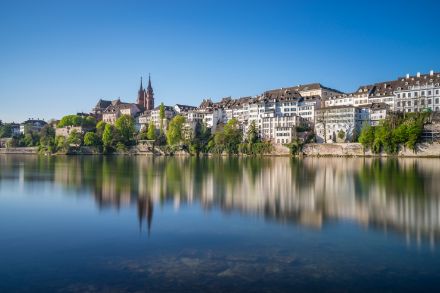 A scenic view of Basel, Switzerland, featuring the historic buildings of Grossbasel along the Rhine River. The calm water reflects the cityscape, including the iconic Basel Minster with its red sandstone towers. The sky is clear and blue, enhancing the vibrant contrast between the lush green trees and the traditional Swiss architecture.
