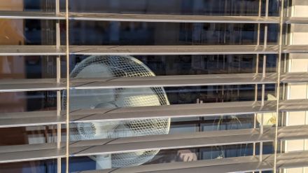 Vue de l’extérieur à travers une fenêtre dont les stores sont à moitié fermés. Dans la pièce, un ventilateur est placé près de la fenêtre.