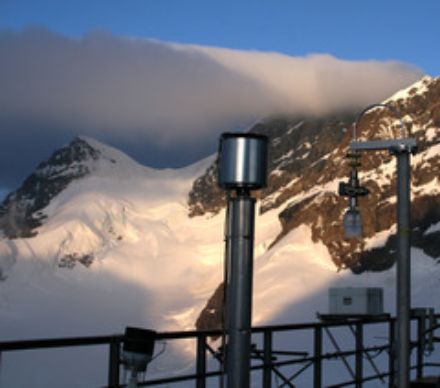 Les prises d'air sont aménagées sur une terrasse en plein air. L'appareil visible au centre de l'image est constitué d'un tube fixé à la verticale et surmonté d'un récipient métallique à travers lequel l'air est aspiré. Des montagnes et des glaciers sont perceptibles au second plan.