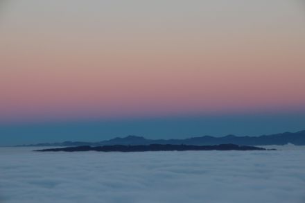 Vue sur une mer de brouillard dont émergent des montagnes. Au fond, une étroite bordure sombre - l'ombre de la Terre - s'étend vers le haut. Au-dessus de celle-ci se détachent des couleurs crépusculaires rougeâtres.