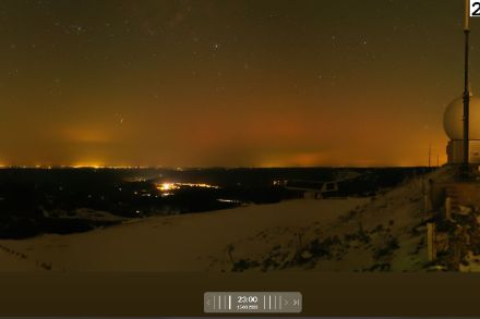 Photo de la webcam de La Dôle prise à minuit en heure locale. Des lueurs rougeâtres apparaissent en direction du nord à travers un fin voile de cirrus. Elles se distinguent nettement de la pollution lumineuse jaune-orangée visible sur l'image. se distingu
