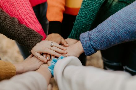 Plusieurs mains se rejoignent pour former un signe de solidarité. Les personnes portent des pulls en tricot colorés, certaines mains sont ornées de bagues. L'image dégage un sentiment de communauté et d'esprit d'équipe.
