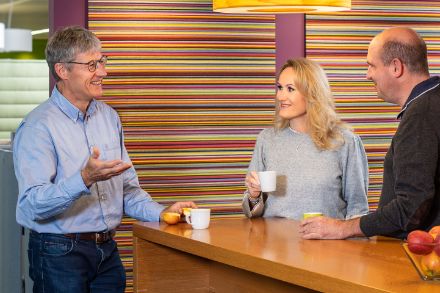 Trois collaborateurs de MétéoSuisse se tiennent près d'une petite cafétéria et boivent un café ensemble. Les deux hommes et la femme discutent entre eux. Sur le comptoir, il y a un récipient avec des pommes. 