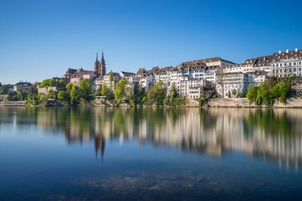 A scenic view of Basel, Switzerland, featuring the historic buildings of Grossbasel along the Rhine River. The calm water reflects the cityscape, including the iconic Basel Minster with its red sandstone towers. The sky is clear and blue, enhancing the vibrant contrast between the lush green trees and the traditional Swiss architecture.
